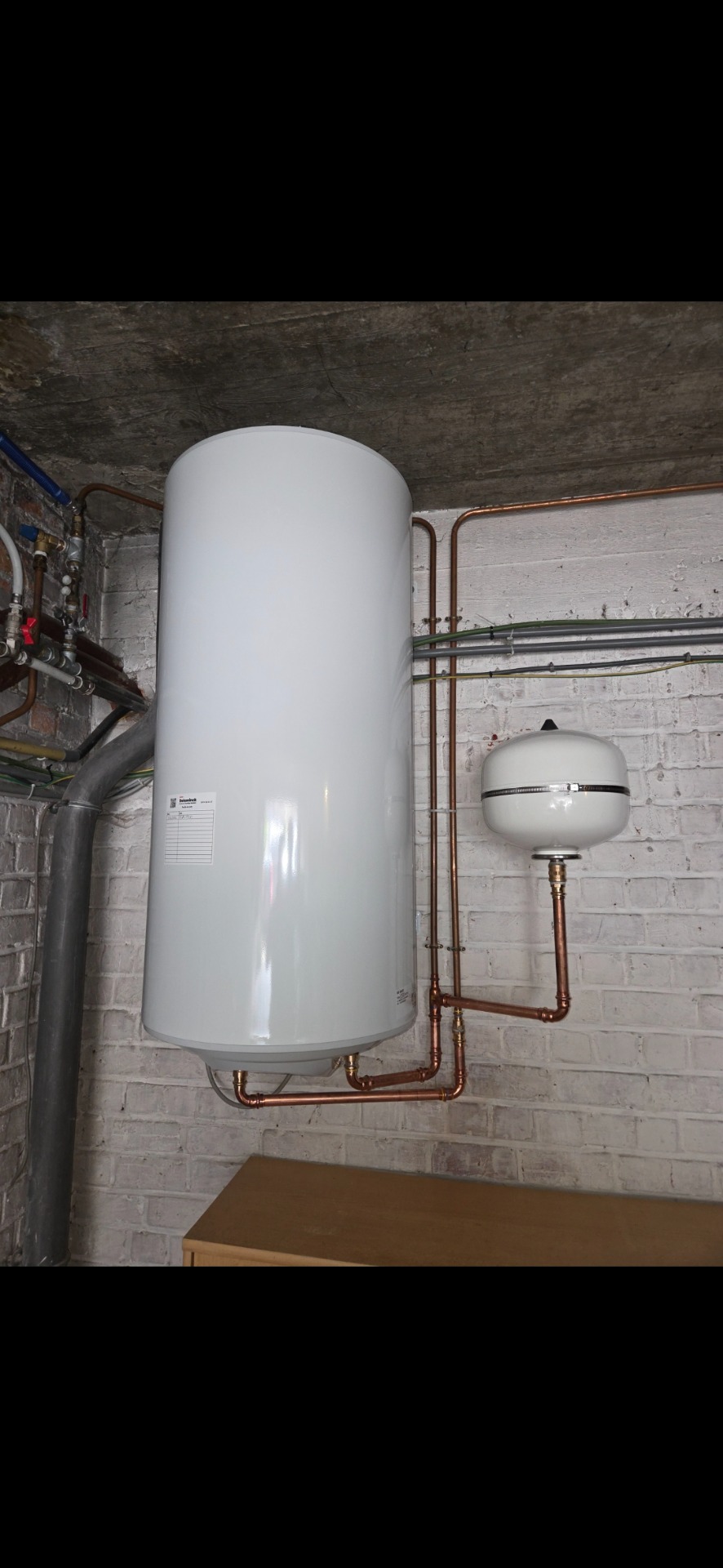 Chauffe-eau blanc vertical et petit ballon, tuyauterie cuivre, mur en briques et conduits techniques.